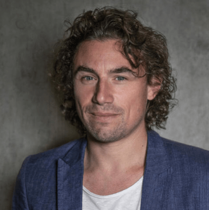 Smiling Frenchman with dark blond curly hair in a white crew neck shirt with a gray-blue blazer in front of a medium gray textured background