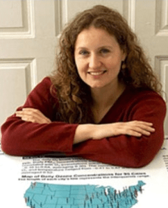 Smiling white woman with long curly auburn hair wearing a red sweater with her arms folded leaning over a table with a map of the usA